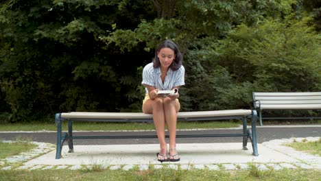 young hispanic woman reading book, sitting with legs crossed on bench at park, wide shot