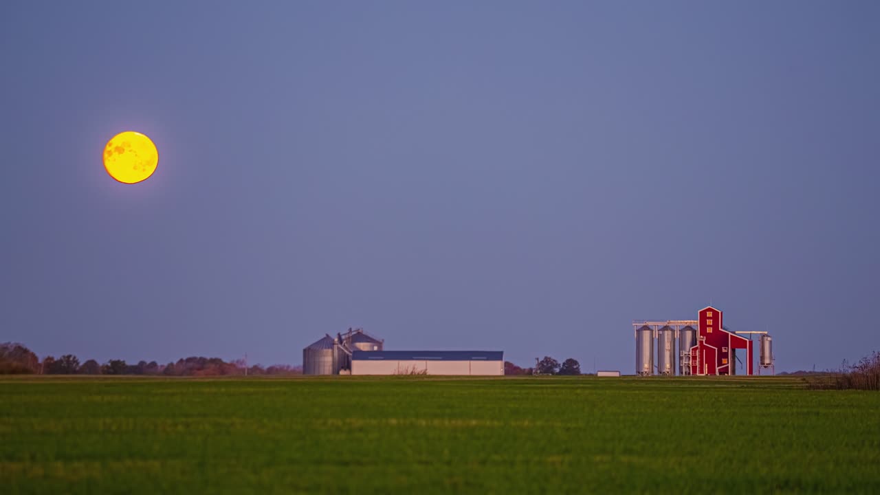 Timelapse: moon shining over a green field with a farm and blue sky, outdoor, static shot