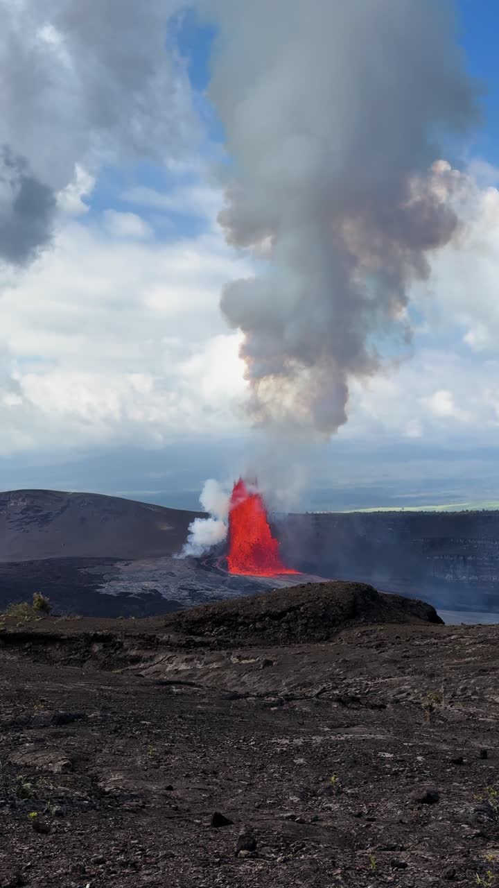 Inquadratura verticale dell'eruzione del Kīlauea sulla Grande Isola delle Hawaii, che mostra una fontana di lava e un'imponente colonna di fumo che si innalza alta nel cielo nuvoloso