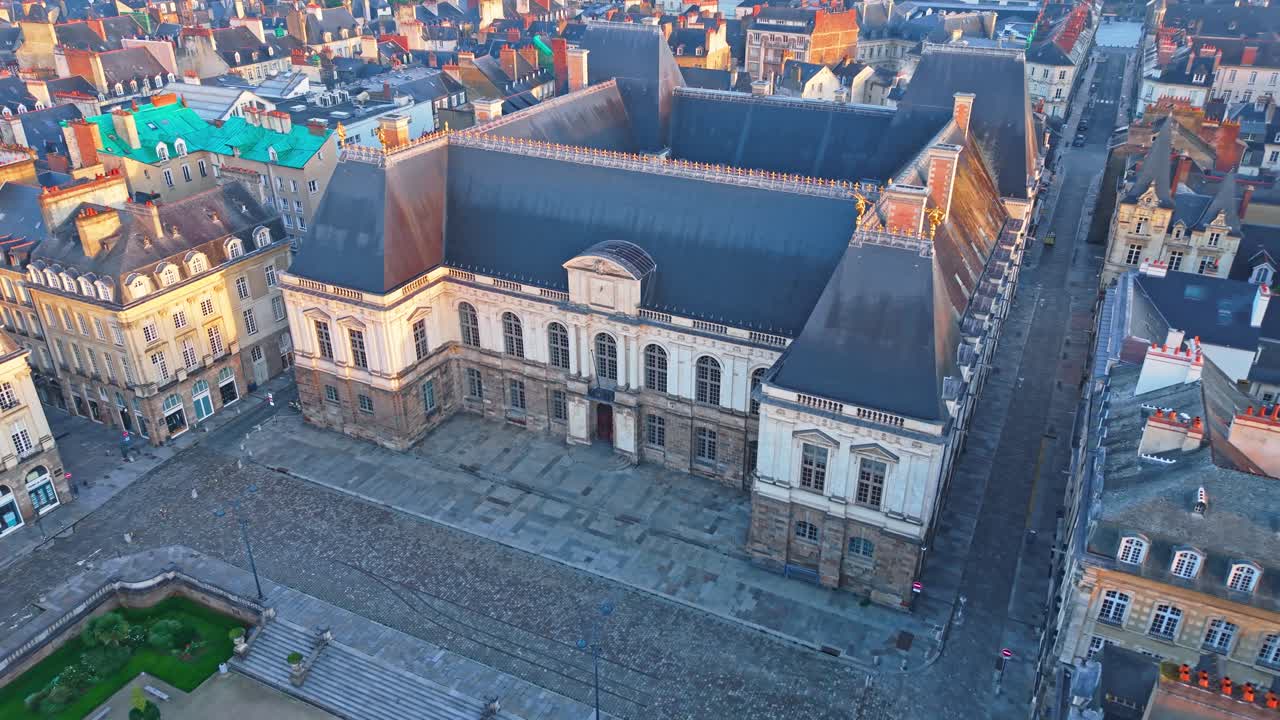 The drone rises and pulls back from the Parlement de Bretagne, revealing the empty square, quiet streets and soft sunrise light over Rennes