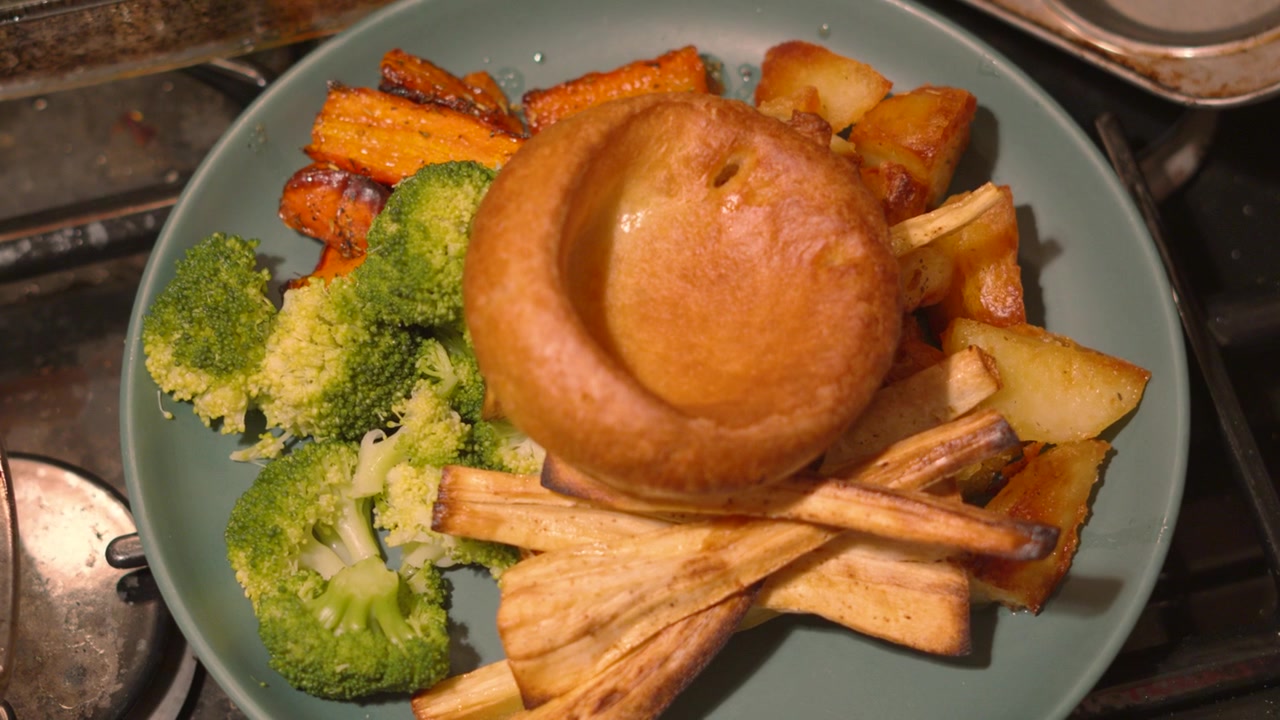 Oversigtsbillede af britisk stegemiddag i køkkenomgivelser med grøntsager, herunder pastinakker, Yorkshire pudding, gulerødder, ovnstegte kartofler og sund broccoli på en tallerken klar til servering 4K