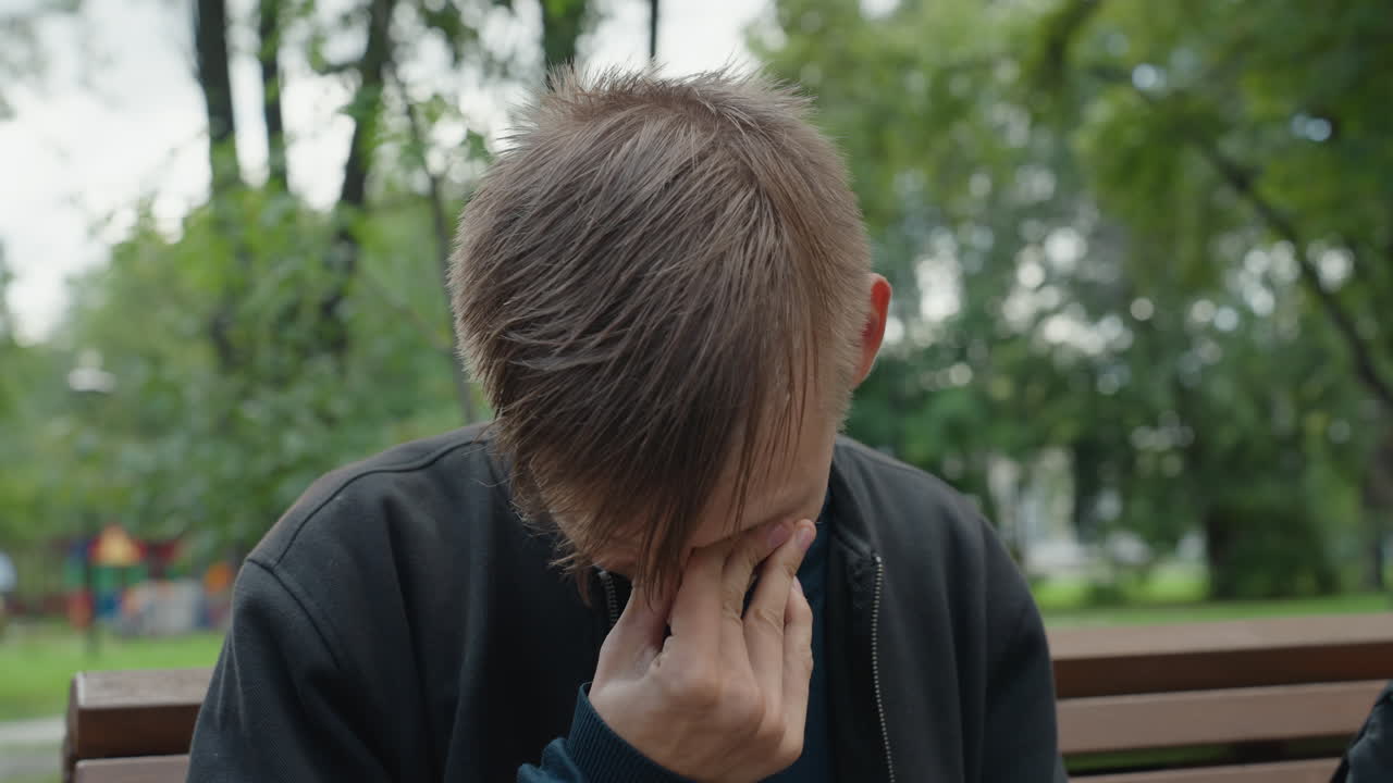 giovane uomo fradicio su una panchina del parco, sottili cambiamenti nella recitazione tra le inquadrature, capelli bagnati e appiccicati alla fronte, sguardo che passa dalla vergogna a una silenziosa risolutezza, illuminazione naturale nuvolosa, alberi e parco giochi sfocato
