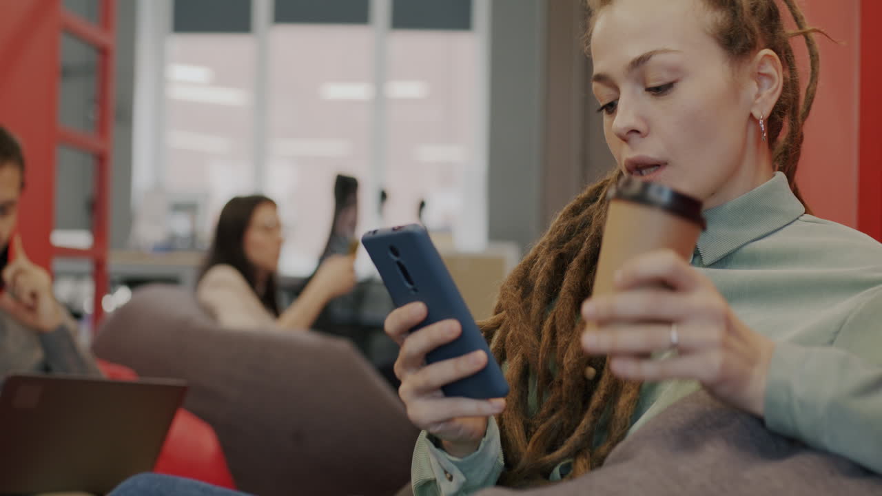 Woman using smartphone and drinking coffee in a coworking space
