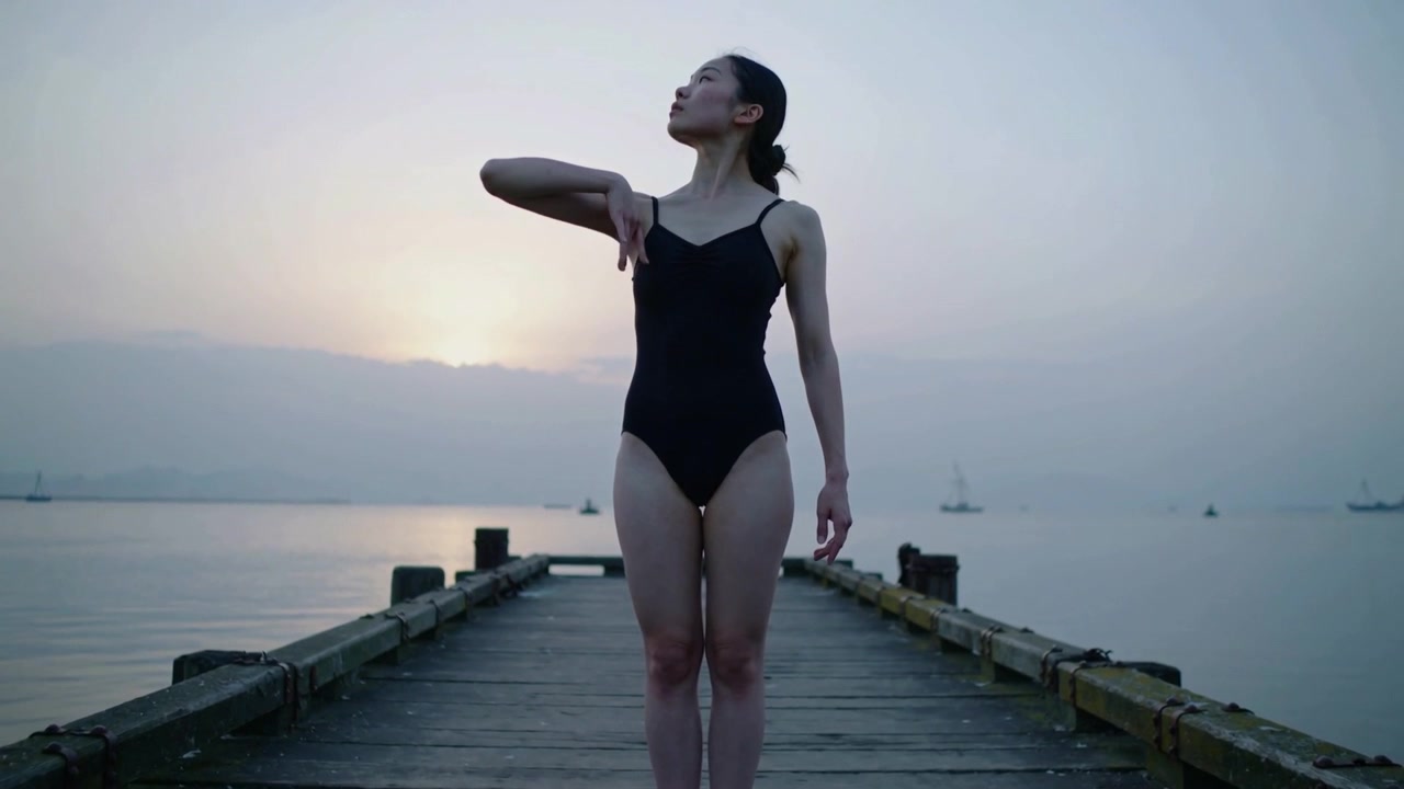 Raising dancer practicing crossing right arm, turning-left on pier with sun low