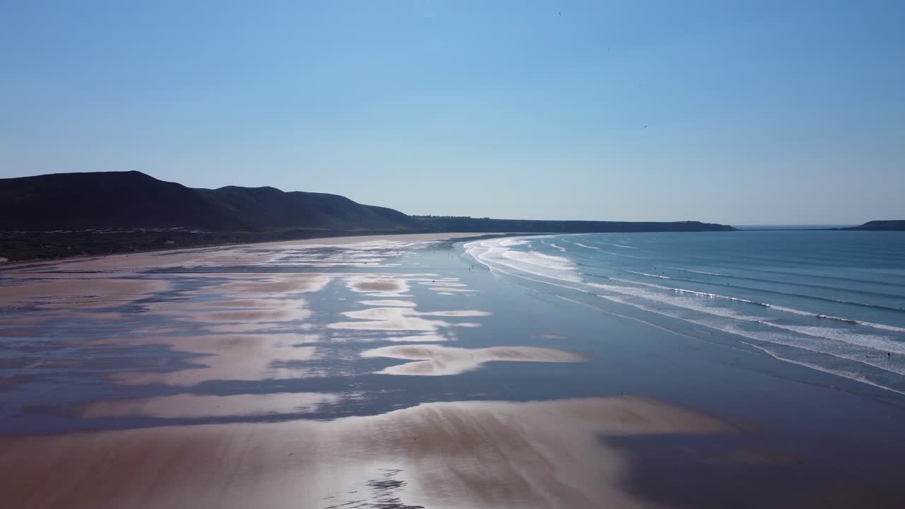 Rising Aerial Over Rhossili Bay with Bright Sun Reflection on Wet Sand at Low Tide with Small Waves and Moorland Silhouette 4K