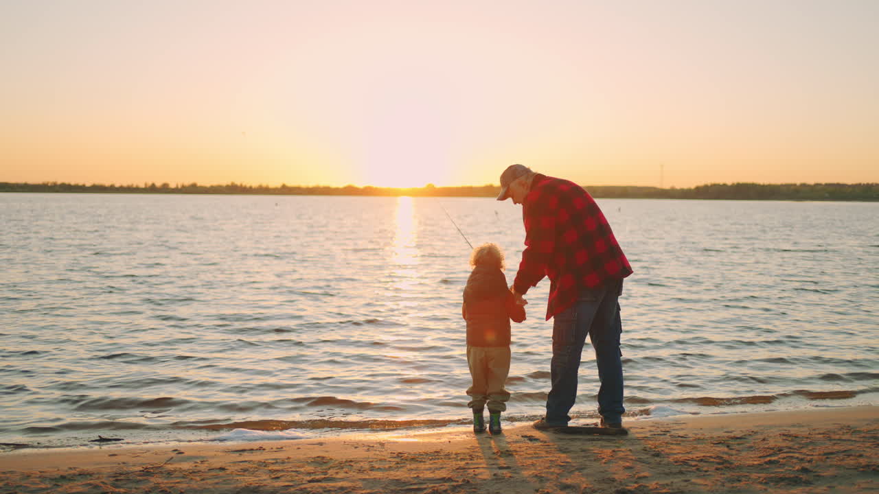 il nonno sta insegnando al suo nipote a pescare con la verga. il vecchio e il bambino sono in piedi sulla riva sabbiosa.