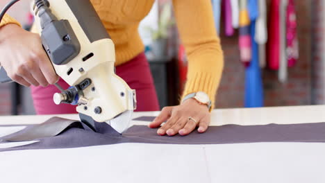 biracial female fashion designer using rotary fabric cutter in studio, slow motion