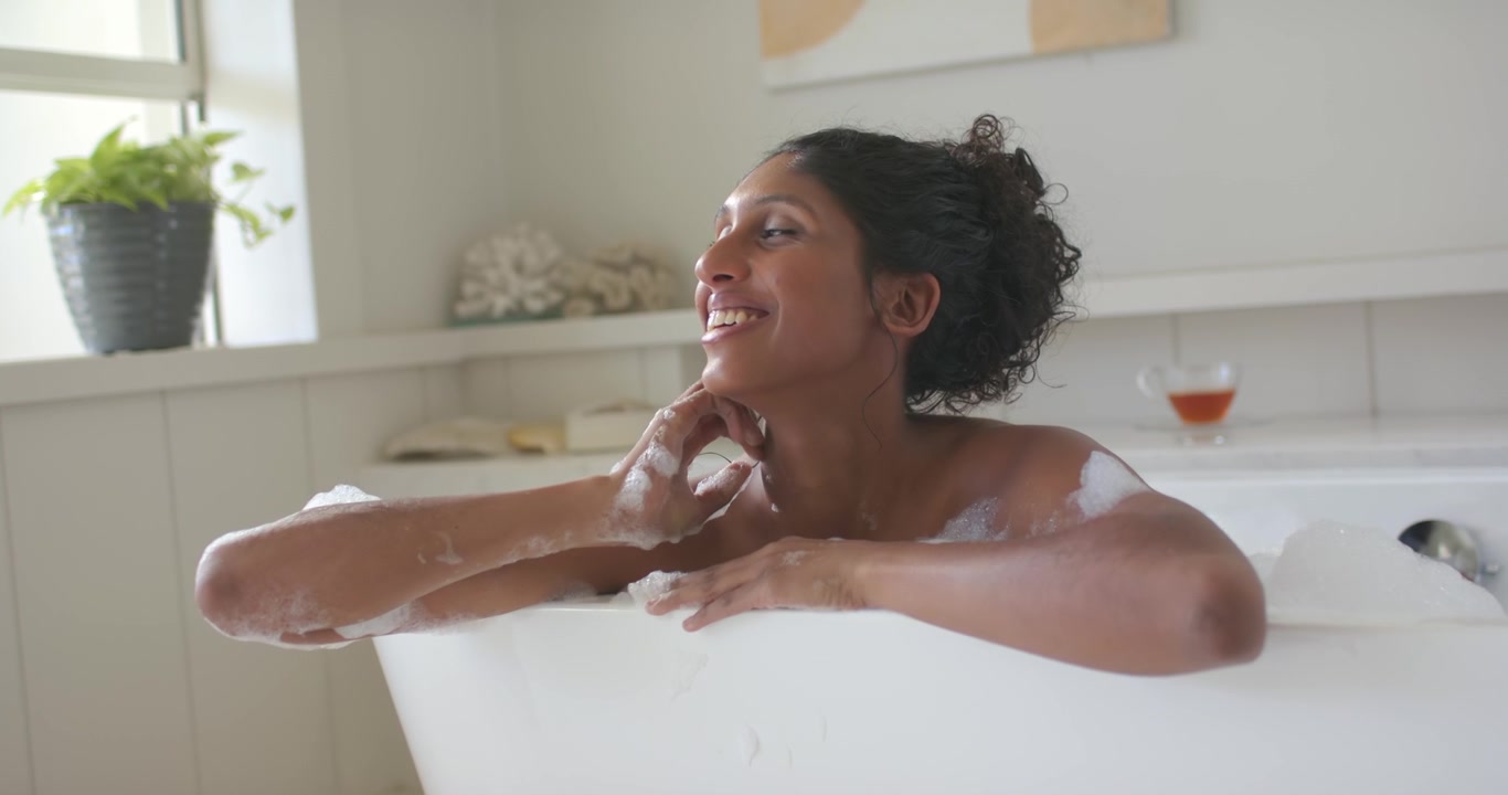 African American woman scooping foam, blowing bubbles, rubbing neck in white tub by window