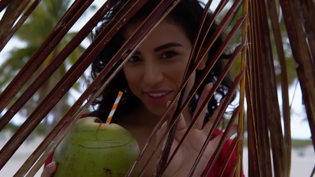 Smiling woman holding green coconut while peeking through the leaves of a palm tree. The beach scene in the background adds to the tropical vibe of the moment