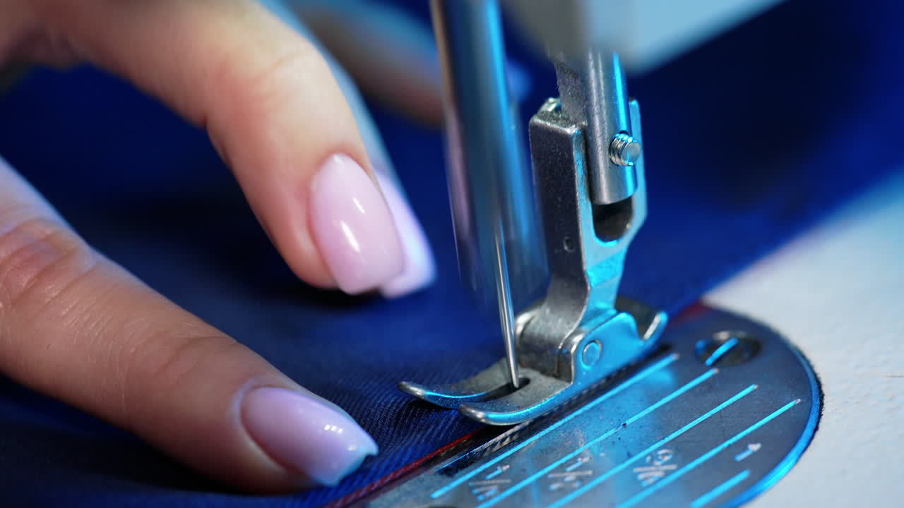 Female hand pushes material through a sewing machine. Sewing machine needle rapidly moves up and down. Tailor sews blue fabric in the sewing workshop. Close-up.