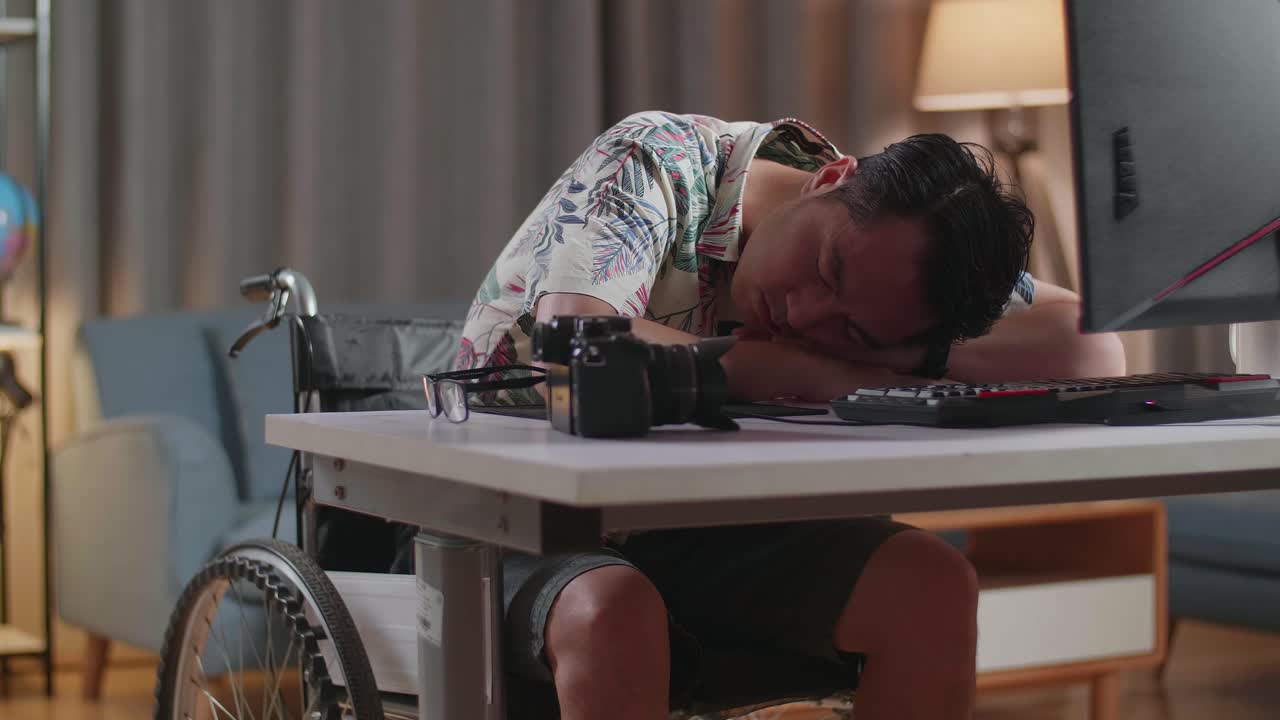 Tired Asian Man In Wheelchair Sleeping While Editing The Video By A Desktop Next To The Camera At Home