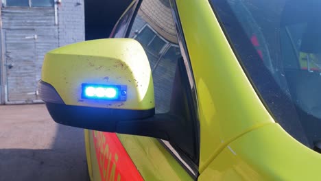 the exterior of yellow paramedic ambulance vehicle with blue flashing warning light in side mirror, sunny day, handheld medium closeup shot