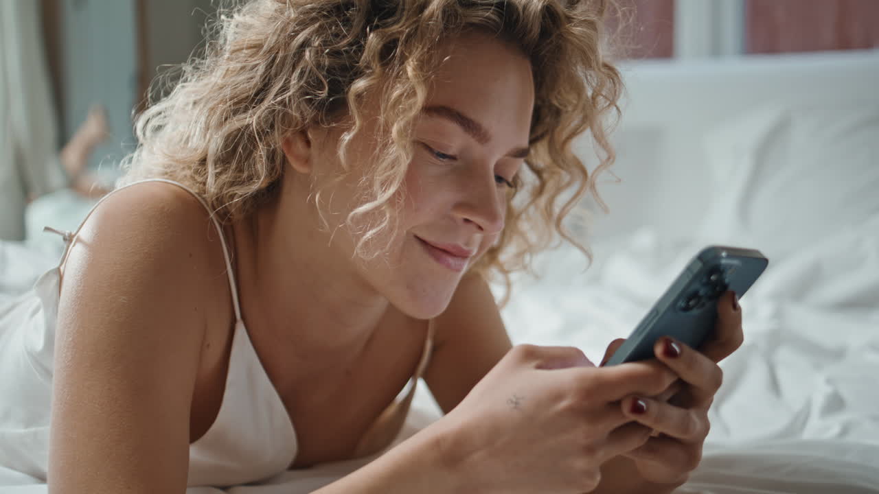 Ragazza allegra che scrive messaggi sullo smartphone nel letto di casa. Primo piano di donna riccia che si gode il web.
