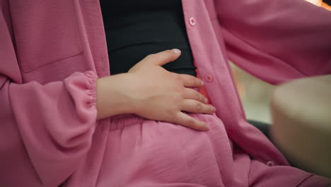 woman wearing a black crop top under a pink dress gently rubbing her stomach with her right hand while the other hand is placed on the table after a meal, showing contentment