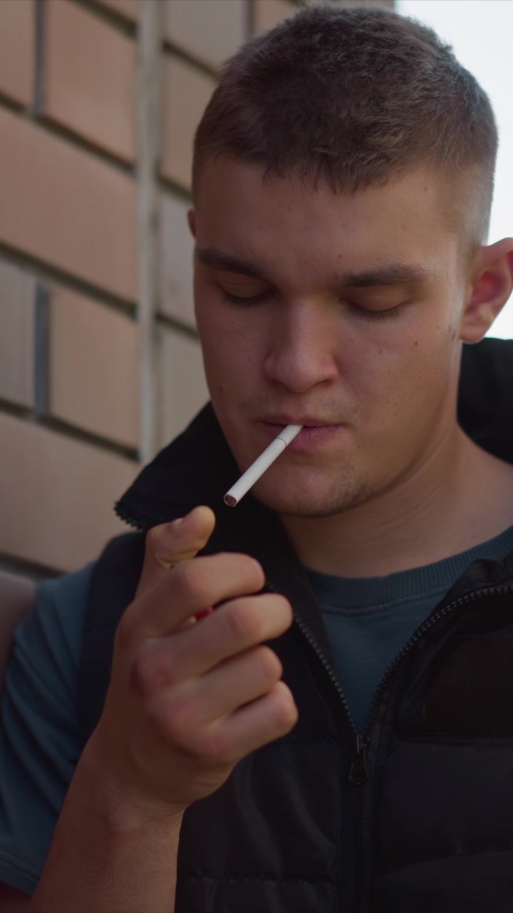 Primer plano de un hombre con chaqueta acolchada negra sosteniendo y fumando un cigarrillo al aire libre, con un edificio de fondo. Gestos de las manos visibles, iluminación de luz diurna suave. Escena centrada en la parte superior del cuerpo del sujeto.