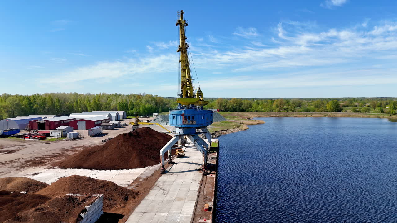 A blue crane stands at a quiet riverside dock with prominent soil mounds and surrounding construction materials, illustrating rural industry and inland cargo operations in a remote setting