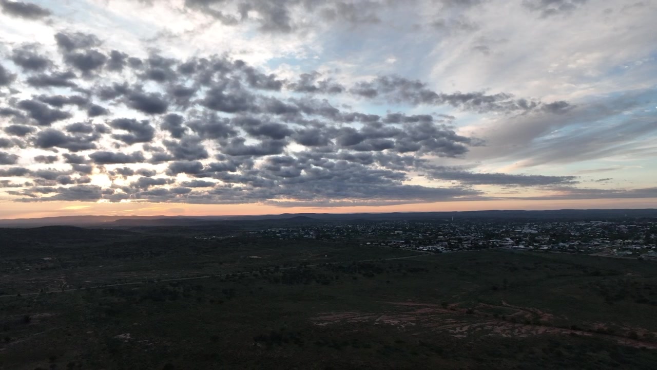 Aerial landscape of Broken Hill beneath long sweeping bands of morning cloud, with subtle orange and pink sunrise colour illuminating the distant horizon.