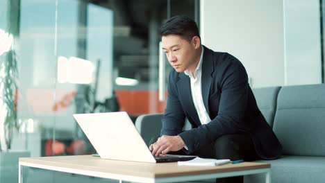 business asian man in suit working on laptop computer in modern office coworking center. confident serious male manager employee satisfied cheerful busy texting,