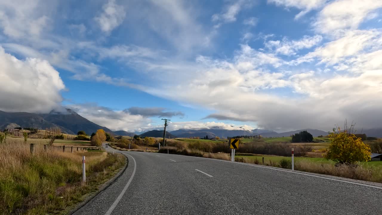 A vehicle travels and stops on a winding rural road through autumn farmland, surrounded by hills and mountains under dramatic, partly cloudy skies with natural daylight
