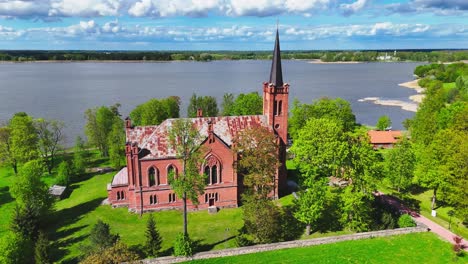 a tall red brick church with a pointed spire overlooks a lakeshore, surrounded by fresh green trees and grassy fields in the lithuanian spring countryside.