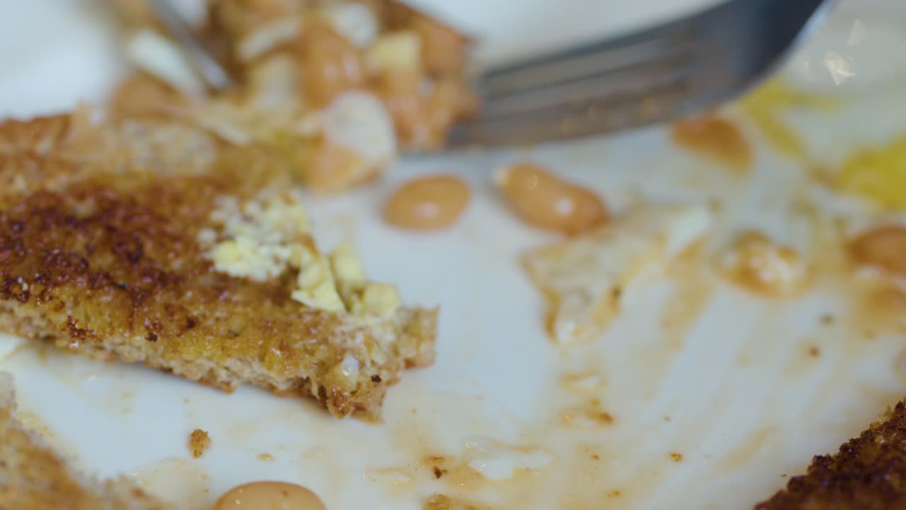 Macro close-up of fork and knife cutting toast, eggs, and beans on a breakfast plate