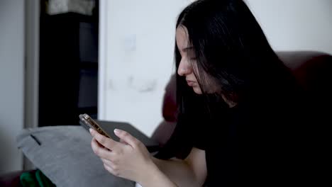 south asian teenage girl browsing the internet on her smartphone while sitting comfortably on a sofa in her cozy living room