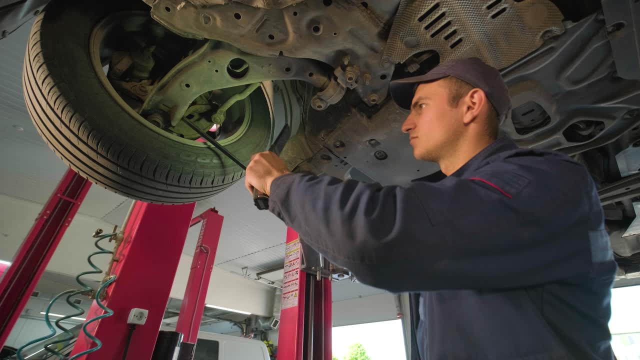 joven mecánico de automóviles en una estación de servicio de reparación inspeccionando la rueda del automóvil y el detalle de la suspensión de un automóvil levantado. vista de abajo.