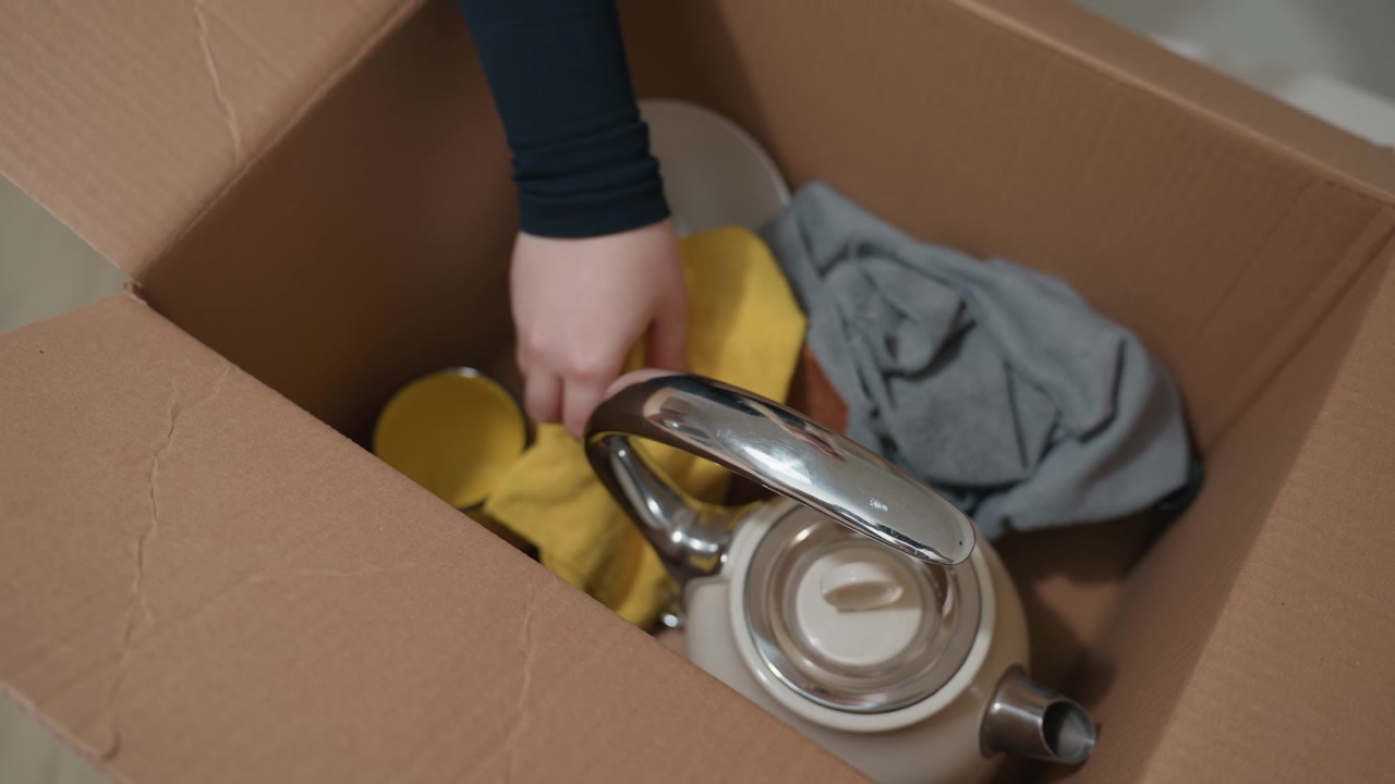 Closeup packing kitchen box fragile items careful renter packing dishware and textiles gentle hands placing towel and ceramic mug into cardboard box soft neutral lighting in new apartment moving day