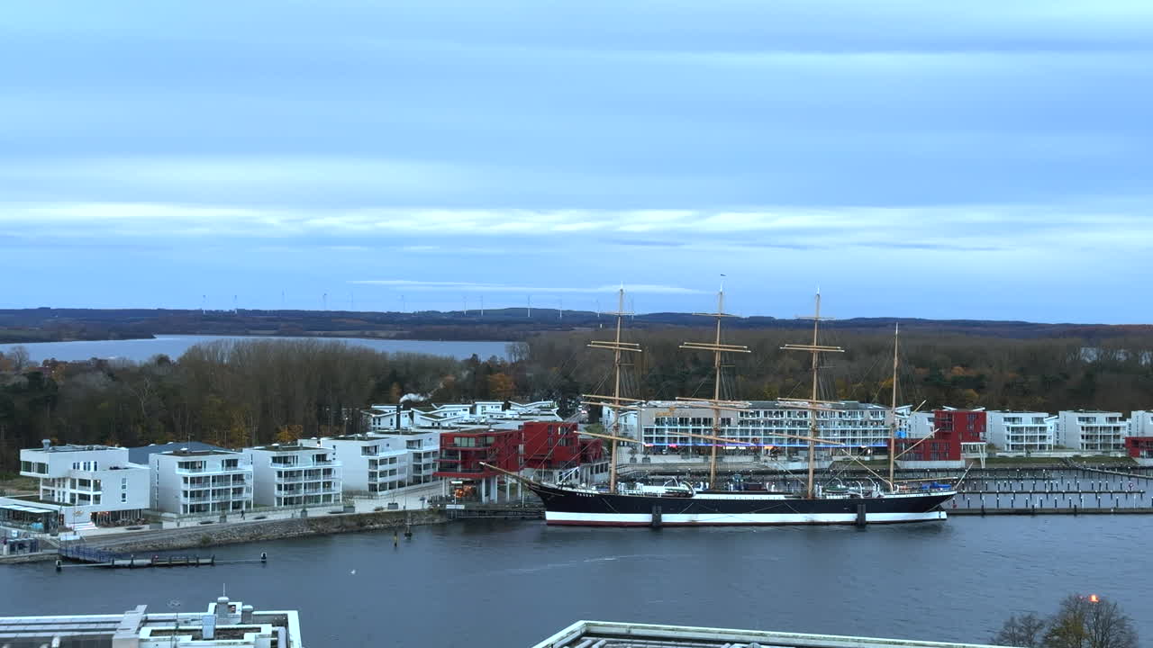 a panoramic view over the waterfront and marina area. A large sailing ship Passat is moored at the pier, standing out prominently against the modern white and red residential buildings nearby