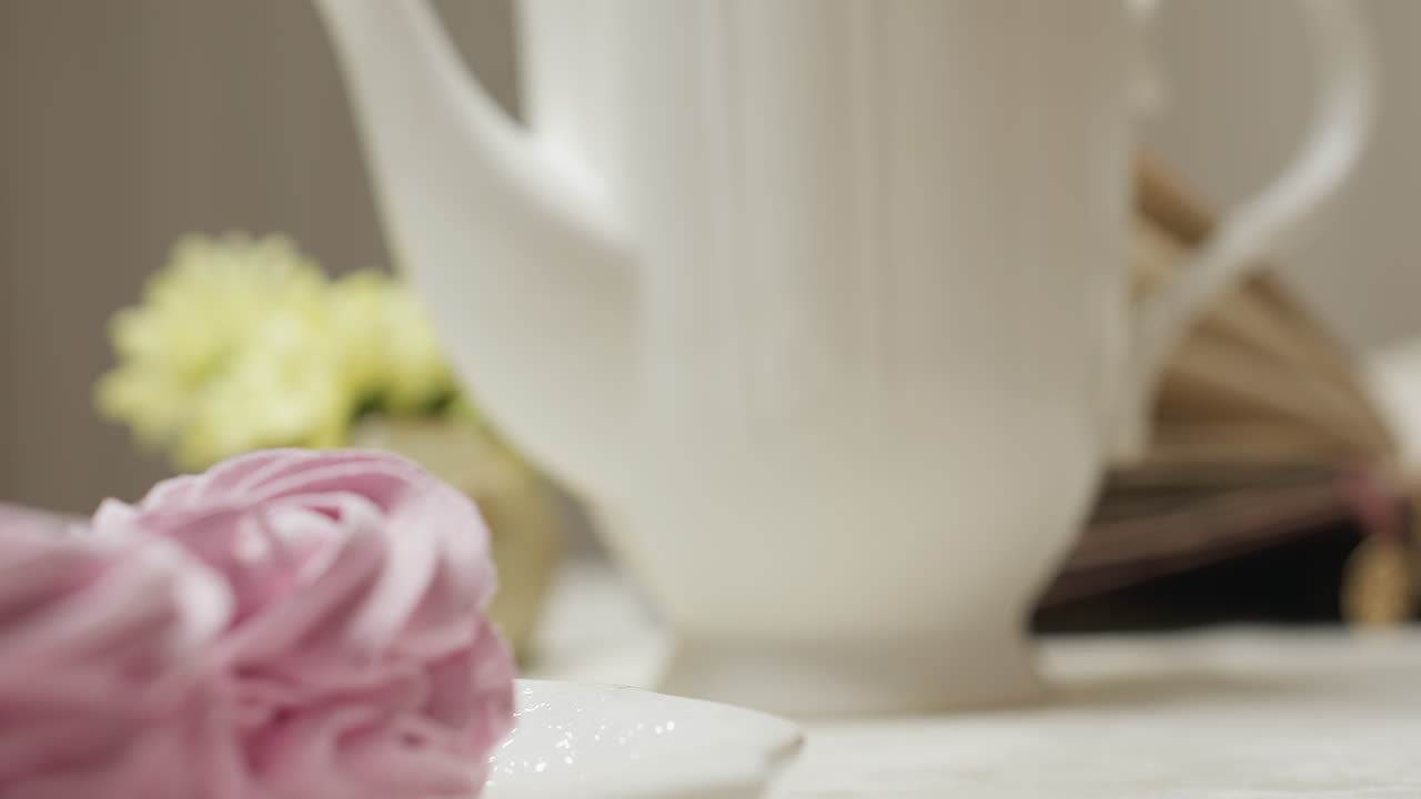 Close up of glass cup filled with herbal tea as person picks it up beside pink cupcake on white plate with soft background of yellow flowers and white teapot in cozy indoor kitchen setting