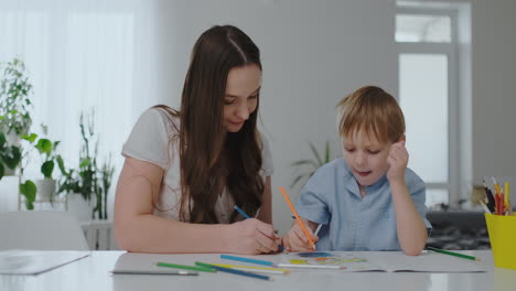 Eine-Junge-Mutter-Sitzt-Am-Tisch-Im-Wohnzimmer-Und-Bringt-Ihrem-Sohn-Bei,-Einen-Bleistift-Zu-Halten-Und-Zu-Zeichnen