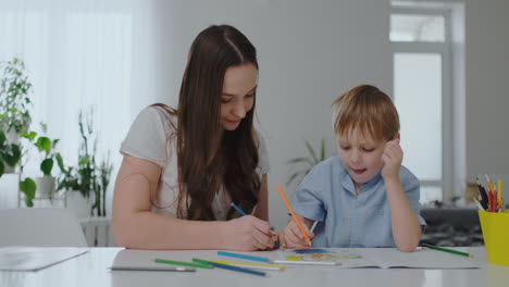 Sentada-En-La-Mesa-Del-Salón,-Una-Joven-Madre-Le-Enseña-A-Su-Hijo-A-Sostener-Un-Lápiz-Y-Le-Enseña-A-Dibujar