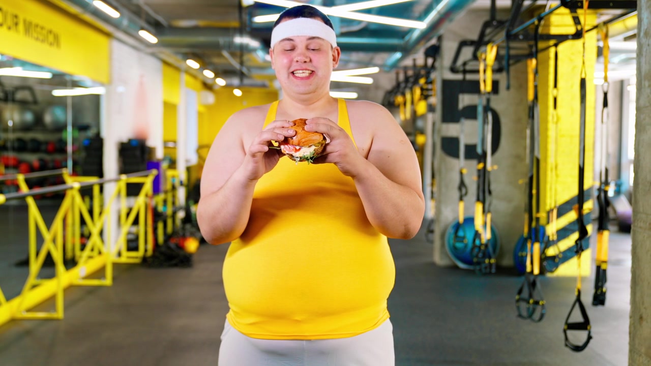 Uomo in sovrappeso che sorride con un hamburger in palestra
