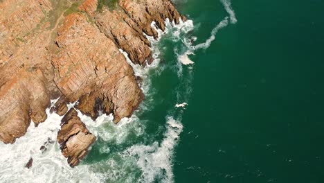 Teal-ocean-waves-breaking-onto-shoreline-with-jagged-rocky-cliff