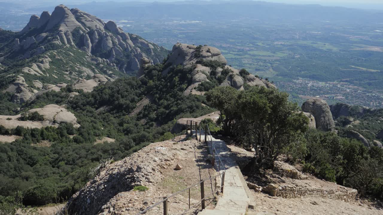 A wide perspective from a rocky mountain ridge at Montserrat, showing a narrow hiking path leading toward the serrated "beehive" conglomerate peaks and the valley below