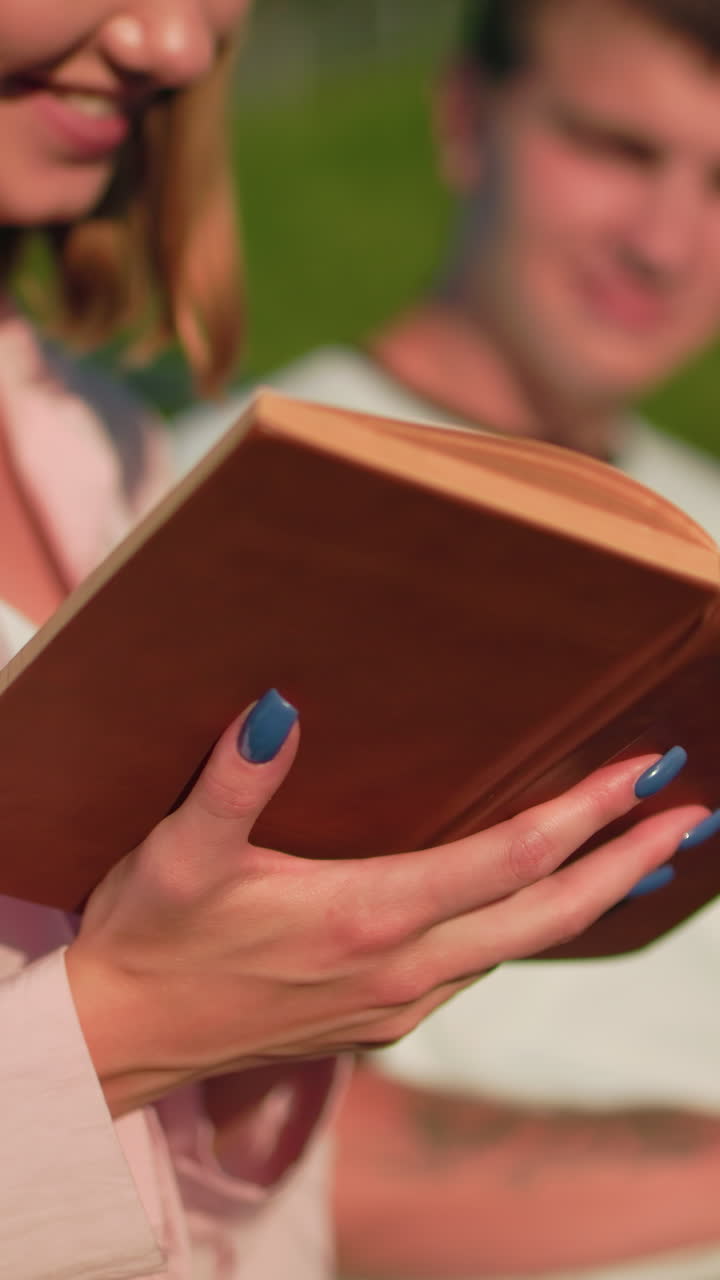 primer plano de la dama de rosa sosteniendo un libro y leyendo mientras su amiga en el fondo escribe en la computadora portátil y asente de acuerdo, el fondo incluye colina verde, banco al aire libre y poste en la cálida luz del sol