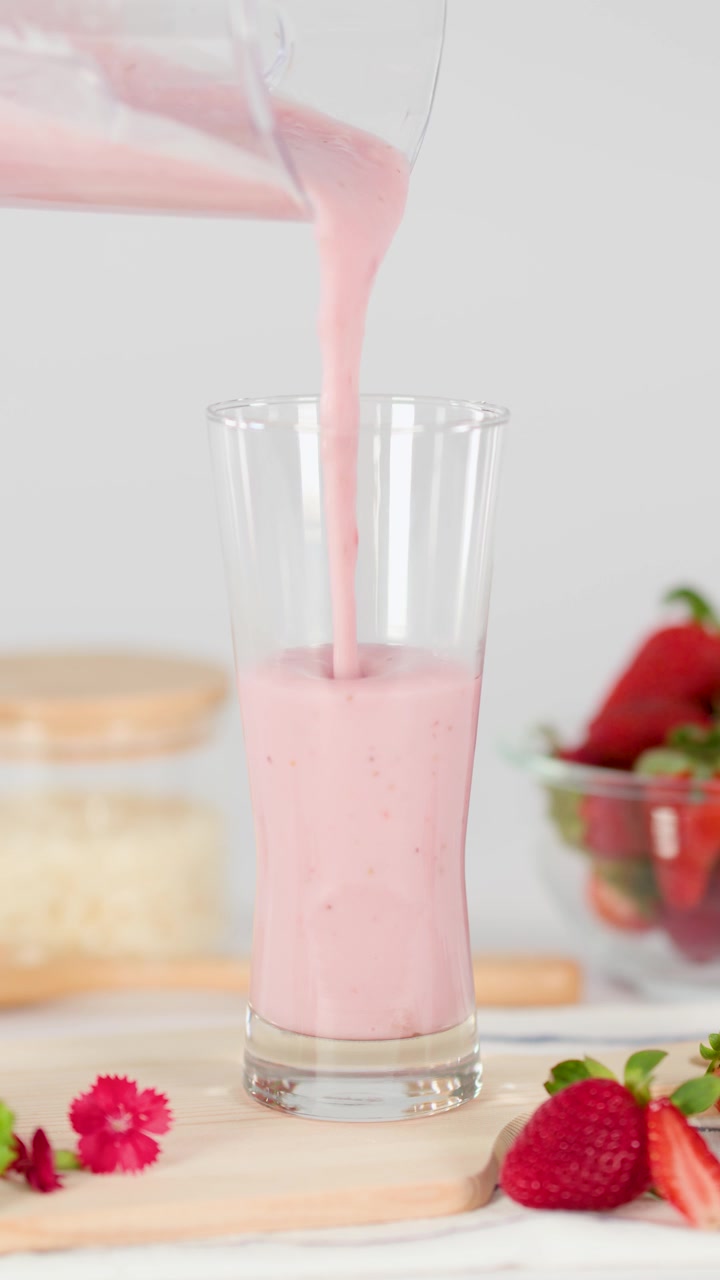 Batido de fresa rosa se vierte suavemente en un vaso, rodeado de fresas frescas, con luz diurna brillante y toma estática.