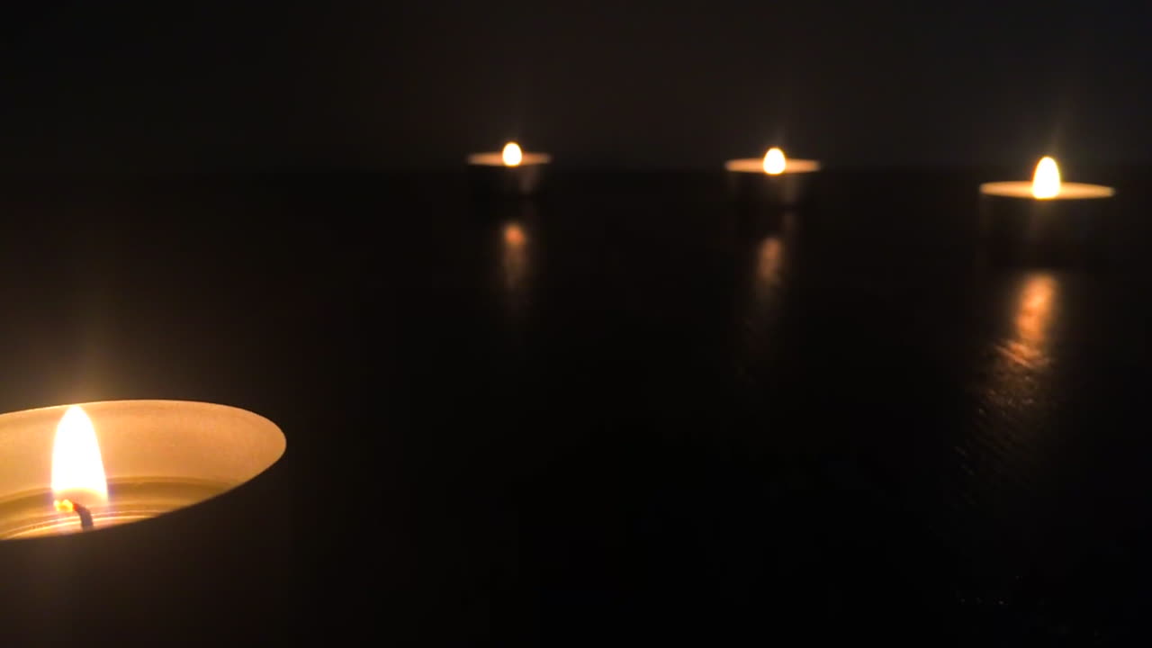 Angled view of four candles with flames and flares on a wooden table in the dark, in slow motion