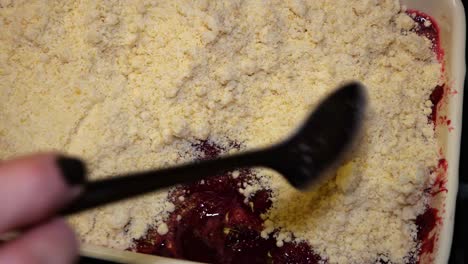 a feminine hand uses a spoon to sprinkle crumble mix on top of a red berry crumble in slow motion