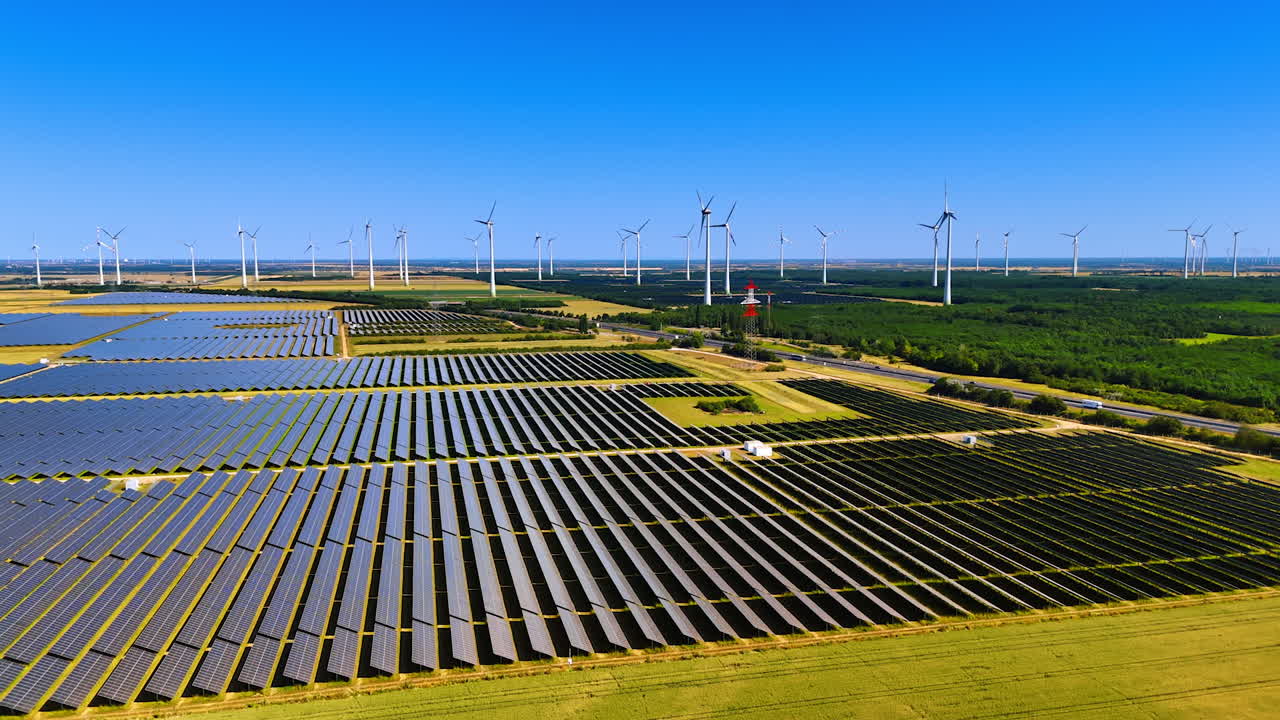 Rows of modern solar panels in the agricultural plantations. Multiple wind mills work at backdrop. Aerial view