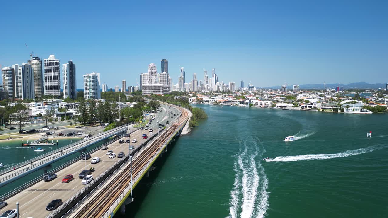 Marine craft travel under a busy road bridge with a towering urban city high-rise in the distance. Drone view