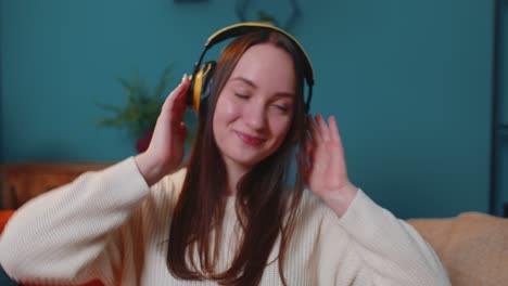 overjoyed young woman in wireless headphones dancing, singing on cozy couch in living room at home