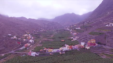 Revealing-aerial-view-of-Hermigua-beach,-valley,-banana-plantation,-colorful-houses-and-mountains-in-hermigua-valley,-North-of-La-Gomera,-Canary-Islands,-Spain-on-a-cloudy-day