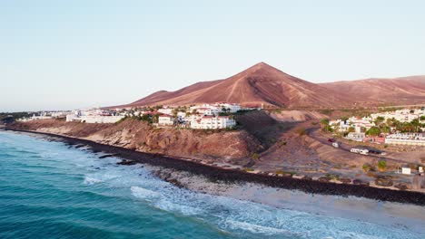 fuerteventura küstenlinie mit vulkangebirge, ozeanwellen und küstenstadt bei sonnenaufgang, luftansicht