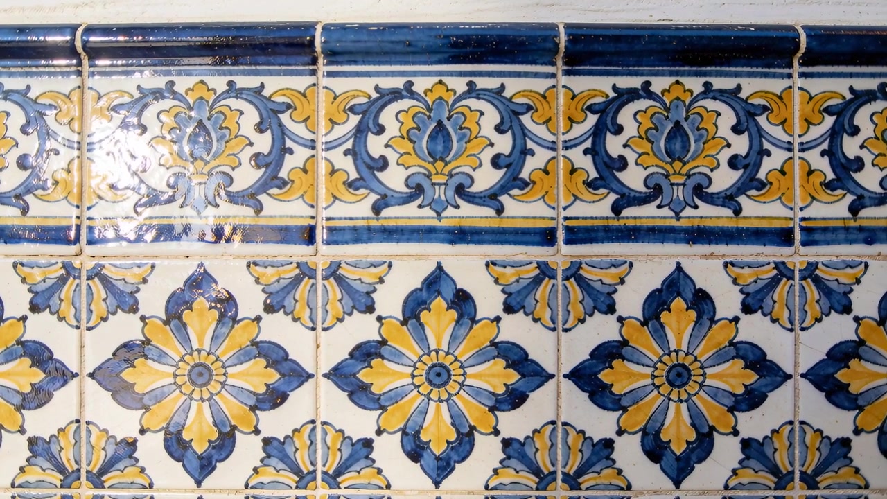 Câmera em movimento panorâmico capturando azulejos esmaltados com rosetas azuis em um corredor, revelando reflexos mutáveis.