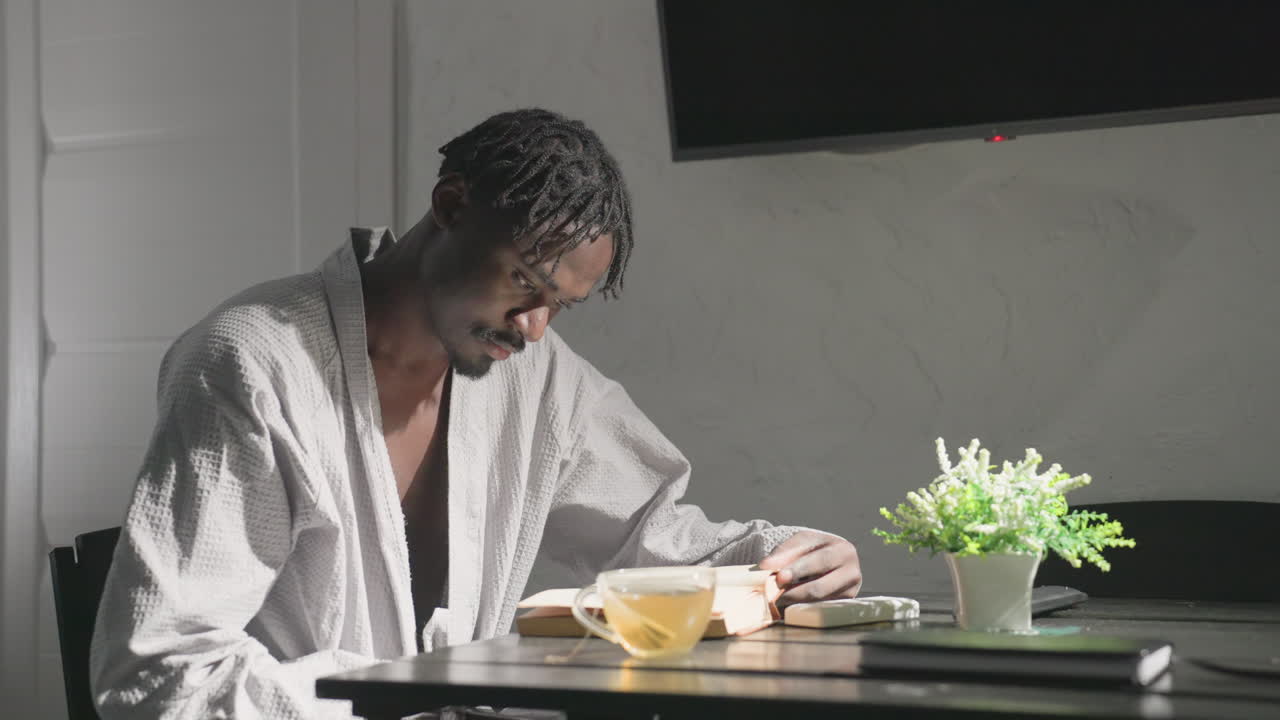 Person in white robe sitting at table in cozy, modern living space sipping tea while reading book. Bright sunlight and soft lighting create calm and relaxed atmosphere perfect for unwinding