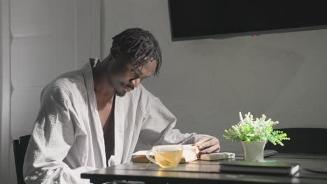 person in white robe sitting at table in cozy, modern living space sipping tea while reading book. bright sunlight and soft lighting create calm and relaxed atmosphere perfect for unwinding