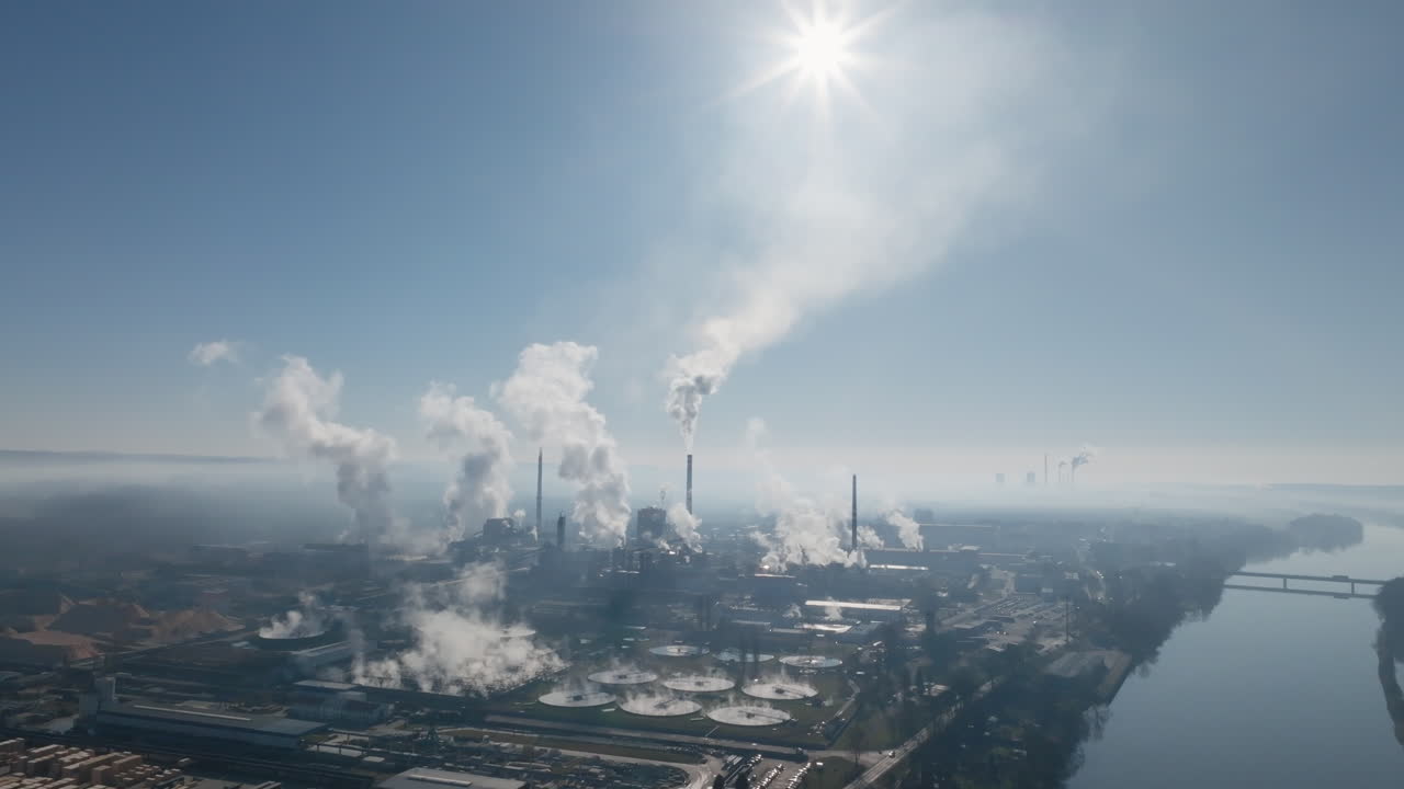 Ampia vista aerea dello skyline della zona industriale di Štětí sotto un forte bagliore solare, con pennacchi di vapore che si alzano da diversi camini e si disperdono all'orizzonte.