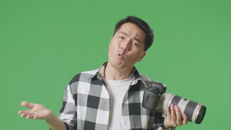 혼란스러운 아시아인 사진작가가, 카메라에서 사진을 보고, 왜라고 말하고, 스튜디오에서 녹색 스크린 배경에 서서 의심스럽게 몸짓을 하고 있다.