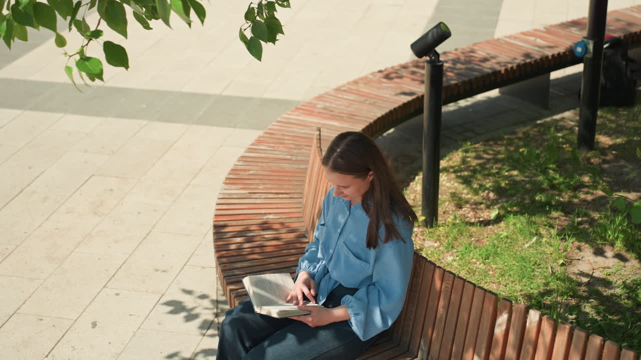 얼룩덜룩한 나뭇잎 아래 곡선형 벤치에 앉아 여행 노트를 적고, 여정과 추억을 기록하며, 카메라 앞에서 자세를 취하고, 따뜻한 햇살과 푸른 주변 환경 속에서 사색에 잠긴 백인 여성